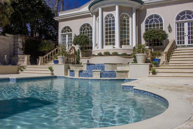 Pool with tiered fountain outside house with large arched windows.
