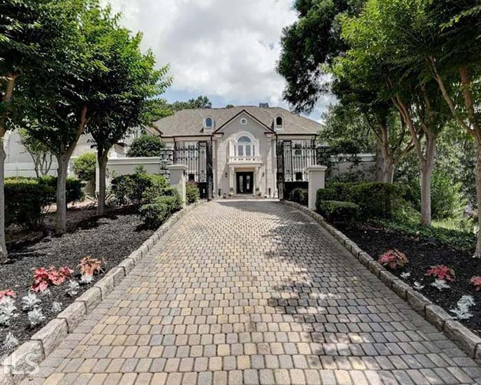 Exterior view of luxury home, looking up cobblestone driveway toward wrought iron gate and front door. Allee of trees and landscaping along both sides of driveway.